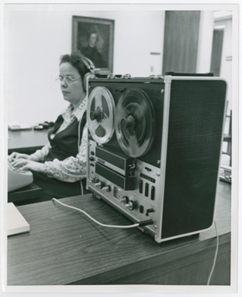 Eine Frau sitzt mit Kopfhörern an einer Schreibmaschine; neben ihr steht ein Tonbandgerät.