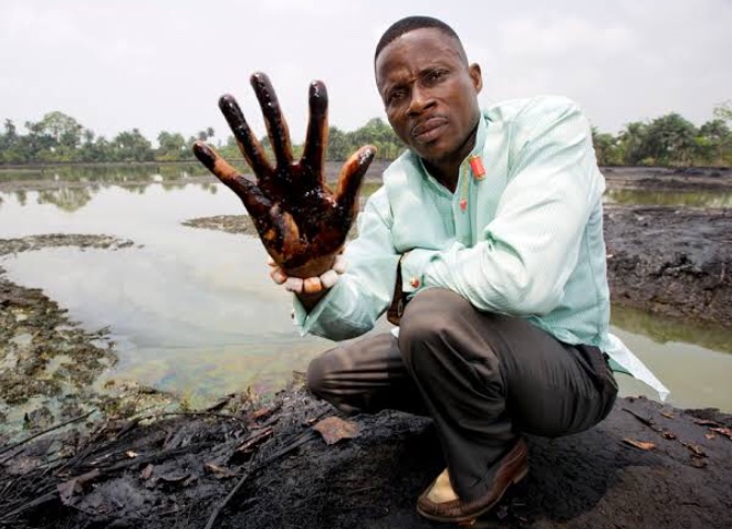 Ein Mann sitzt inmitten eines Ölfeldes und hält seine ölverschmierte Hand hoch.