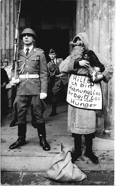 Ein Mann steht neben einem Wachsoldaten am Brandenburger Tor mit einem Schild, auf dem steht: Hilfe! Ich bin wohnungslos, arbeitslos und habe Hunger