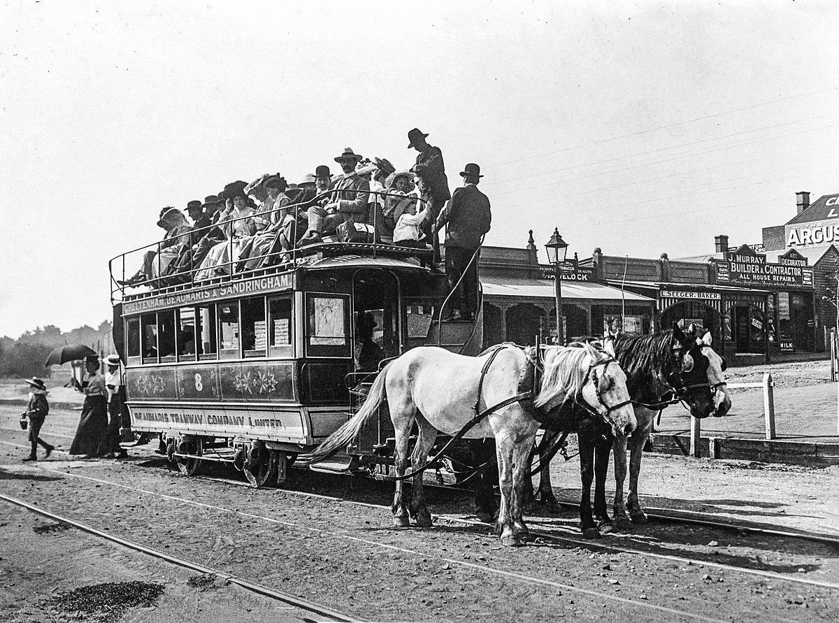 Ein Wagen, auf dem oben Menschen im Freien sitzen, wird von zwei Pferden gezogen.