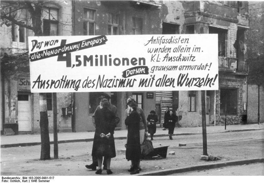 Schwarz-Weiß-Aufnahme eines Transparentes mit der Forderung nach der Ausrottung des Nazismus. Im Hintergrund sind eine Straße, mehrere Menschen und teilweise zerstörte Häuser zu sehen.