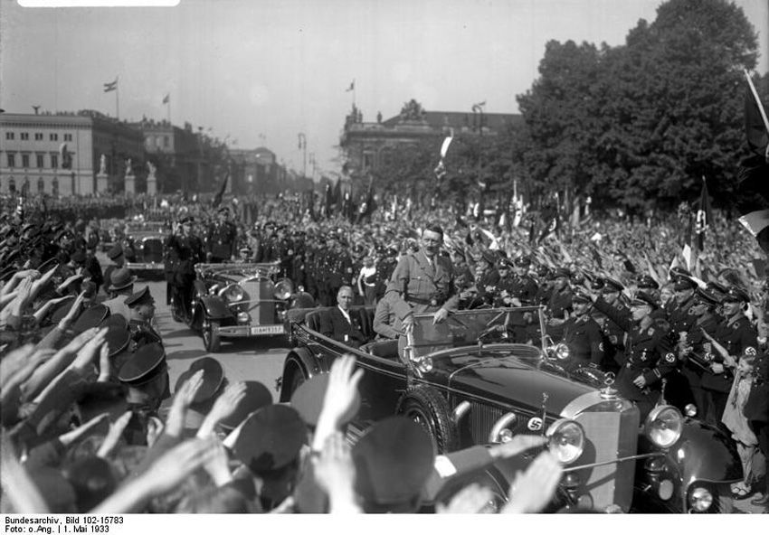 Schwarz-Weiß-Fotografie von Adolf Hitler in einem offenen Wagen während einer Parade, umgeben von einer jubelnden Menschenmenge mit erhobenem Hitlergruß.