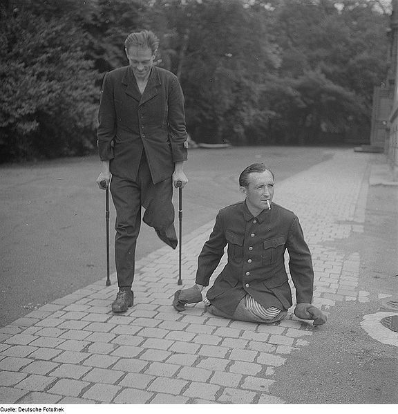 Schwarz-Weiß-Fotografie zweier Männer auf einem Gehweg. Einer mit Krücken und einem amputierten Bein, der andere mit beiden Beinem amputiert daneben am rauchen.
