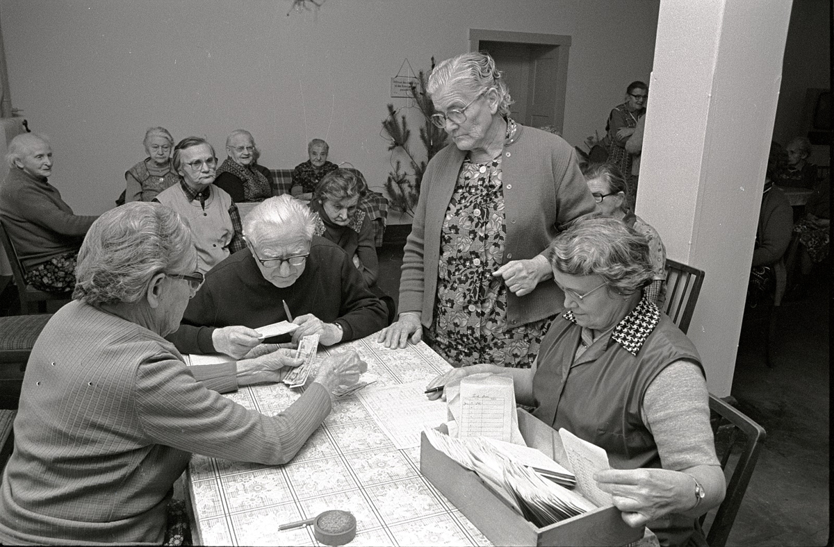 Schwarz-weiß Foto älterer enschen die um einen Tisch mit vielen Zetteln sitzen. Im Hintergrund weitere alte Menschen