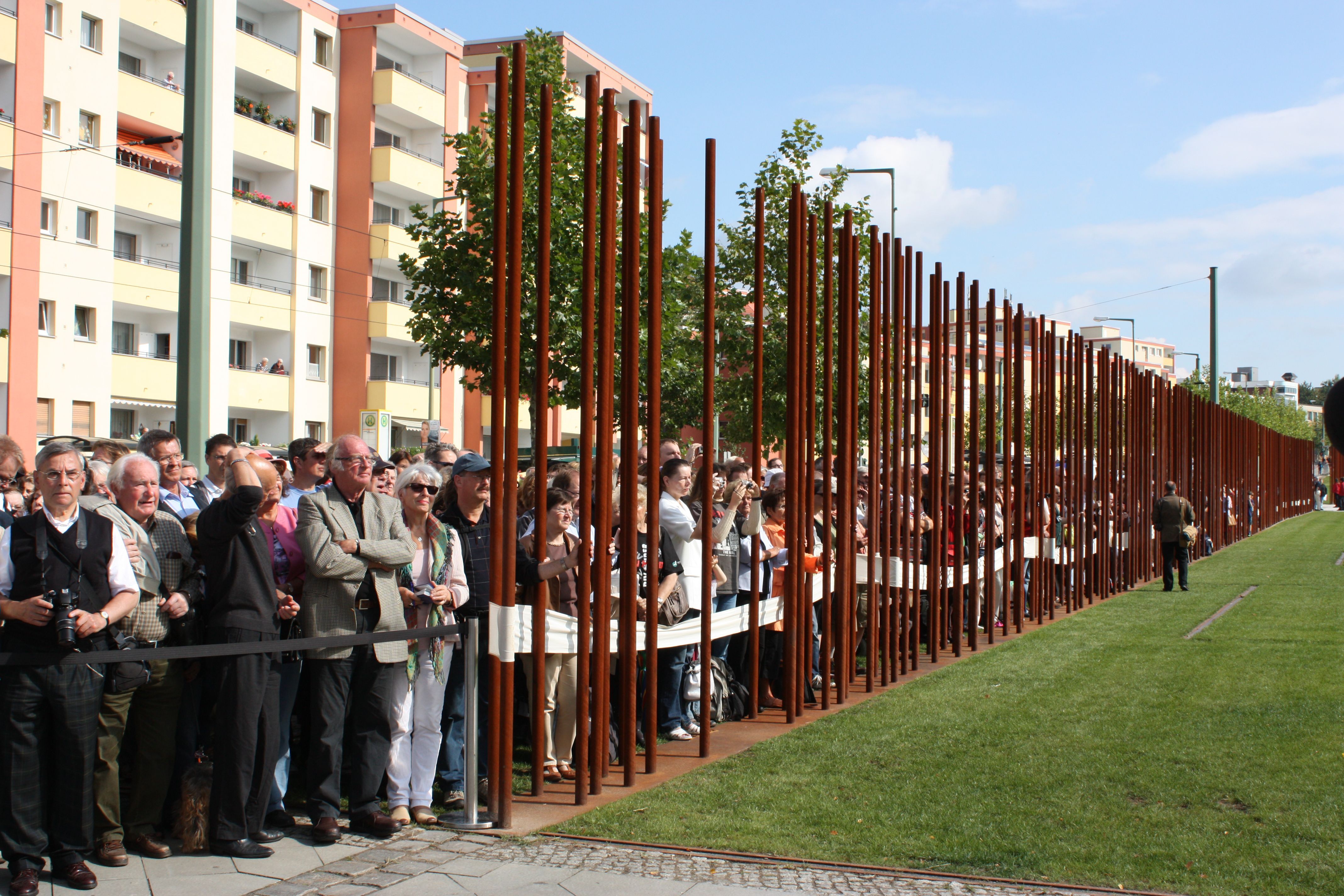 Foto einer Menschenmenge hinter einer Absperrung aufgereiht. Auf der anderen Seite eine Wiese. Getrennt durch eine Linie an vielen Metallstäbe.