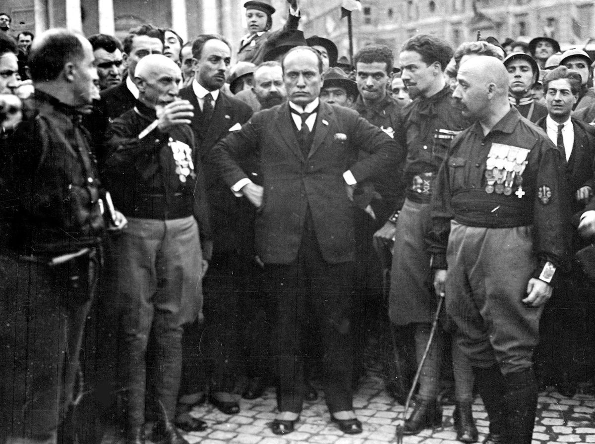 Schwarz-Weiß Fotografie eines grimmig schauenden Mannes mit den Händen in die Hüften gestemmt. Um ihn herum, ihm zugewandt, viele weitere Männer in Uniform, manche mit Abzeichen an der Brust.