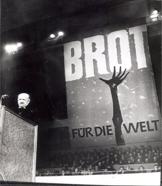 Schwarz-Weiß-Fotografie eines Mannes am Rednerpult. Im Hintergrund Menschen die auf einem Podium sitzen. Über ihnen ein großes Banner mit einer knochigen Hand und der Aufschrift: Brot für die Welt.