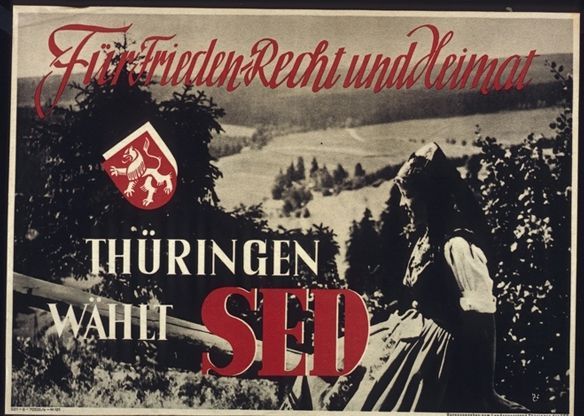 Schwarz-Weiß-Foto einer Frau in Tracht. Im Hintergrund weite Felder. Auf dem Foto weiße und rote Schrift: Für Frieden, Recht und Heimat.Tühringen wählt SED