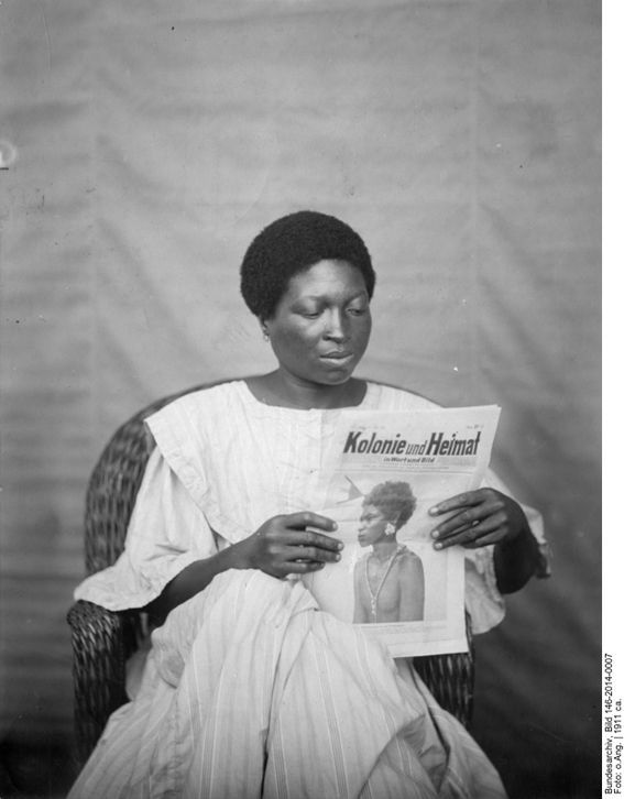 Schwarz-Weiß-Fotografie einer schwarzen Frau in weißem Kleid. Die sitzt auf einem Sessel und liest eine Zeitung mit der Aufschrift: Kolonie und Heimat