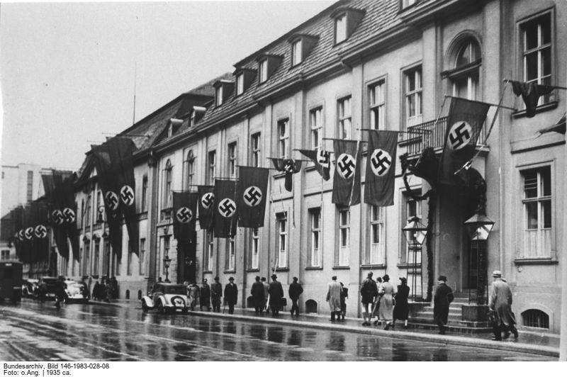 Schwarz-weiß Foto einer Häußerzeile mit vielen heraushängenden Hakenkreuzfahnen. Auf dem Gehweg davor vorrübergehende Menschen.