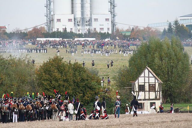Fotografie einer Veranstaltung auf einer großen Wiese. Viele Menschen in Uniformen bewegen sich auf der Wiese. Im Hintergrund ein großes Industrielles Gebäude. Im Vordergrund Bäume und eine kleine Fachwerkhütte.