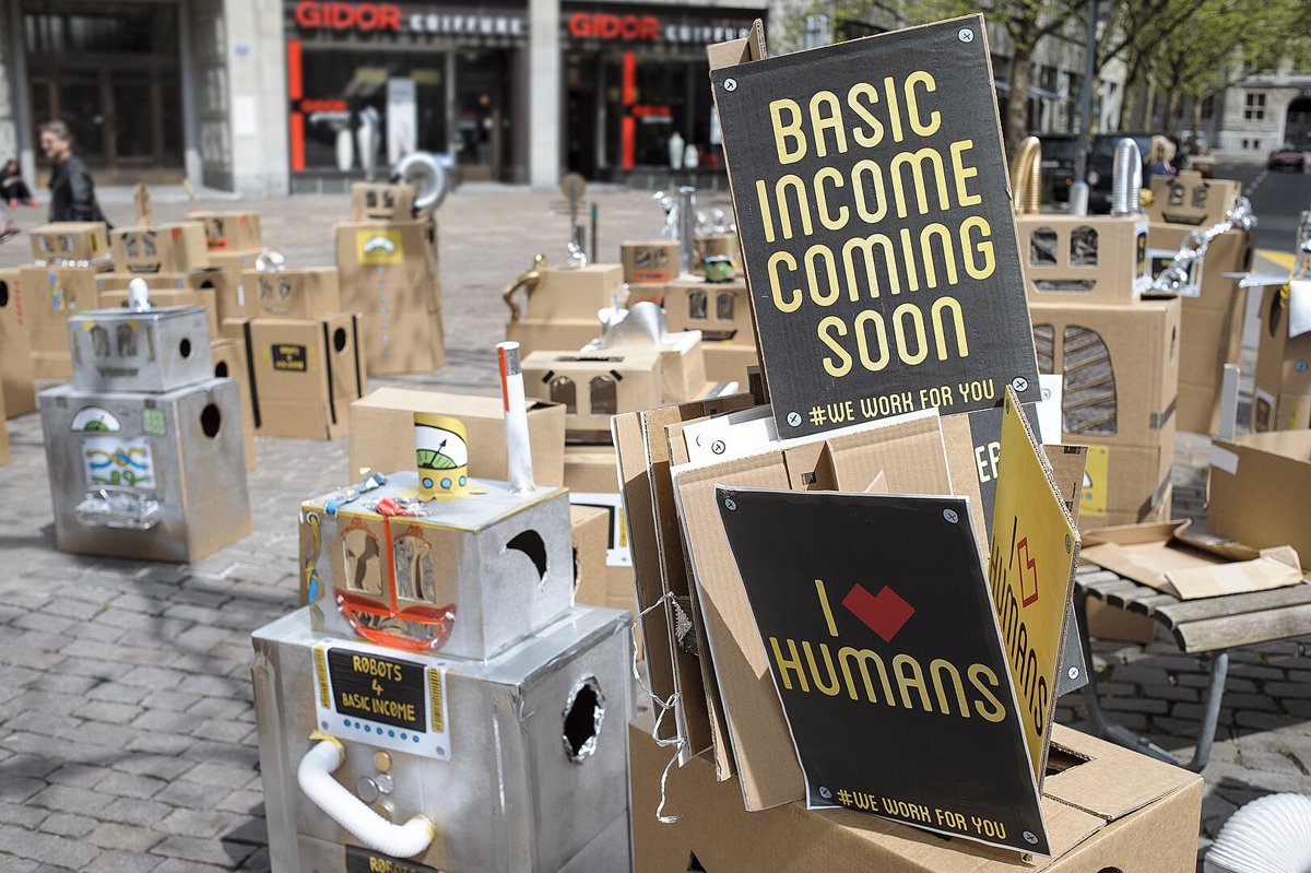 Foto einer Straße voller Pappboxen aus denen Robotter gebastelt wurden. Im Vordergrund ein Schild mit der Aufschrift: basic income coming soon # we worke for you.