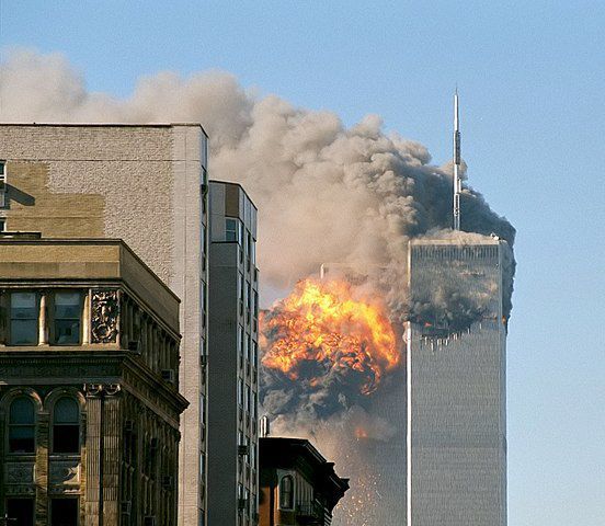 Fotografie einer großen Explosion in einem Hochhaus. Ein zweites Hochhaus daneben ebenfalls am brennen von einer großen Rauchwolke umgeben. Im Vordergrund eine Häußerzeile. 