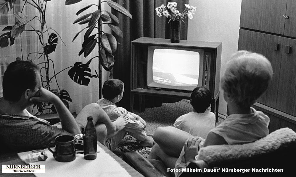 Schwarz-Weiß-Fotografie einer Familie von hinten. Zwei Erwachsene und zwei Kinder sitzen in ihrem Wohnzimmer vor dem laufenden Fernseher.